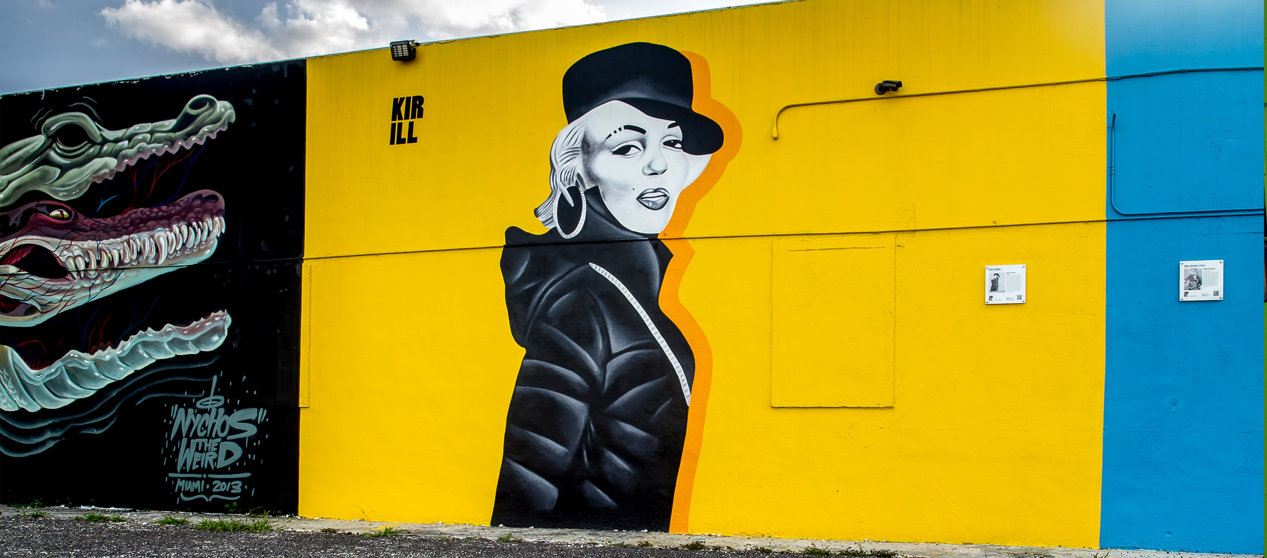Vibrant mural: black-and-white portrait of a woman in a hat on a yellow wall, with a blue panel to the right.