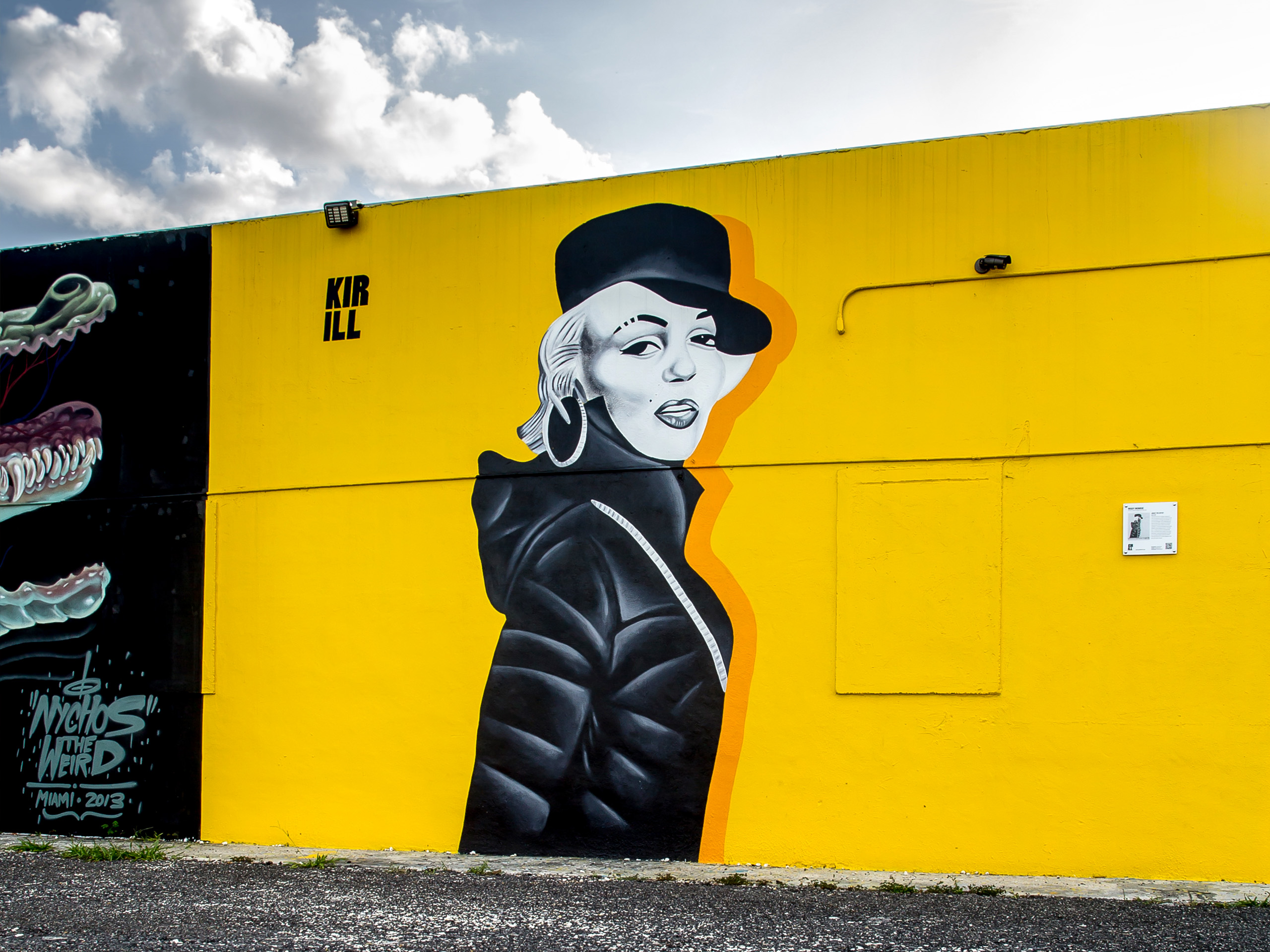 Wall mural on a yellow barrier showing a stylized black-and-white portrait of a person wearing a top hat, with an orange outline, set against a bright blue sky with clouds above.
