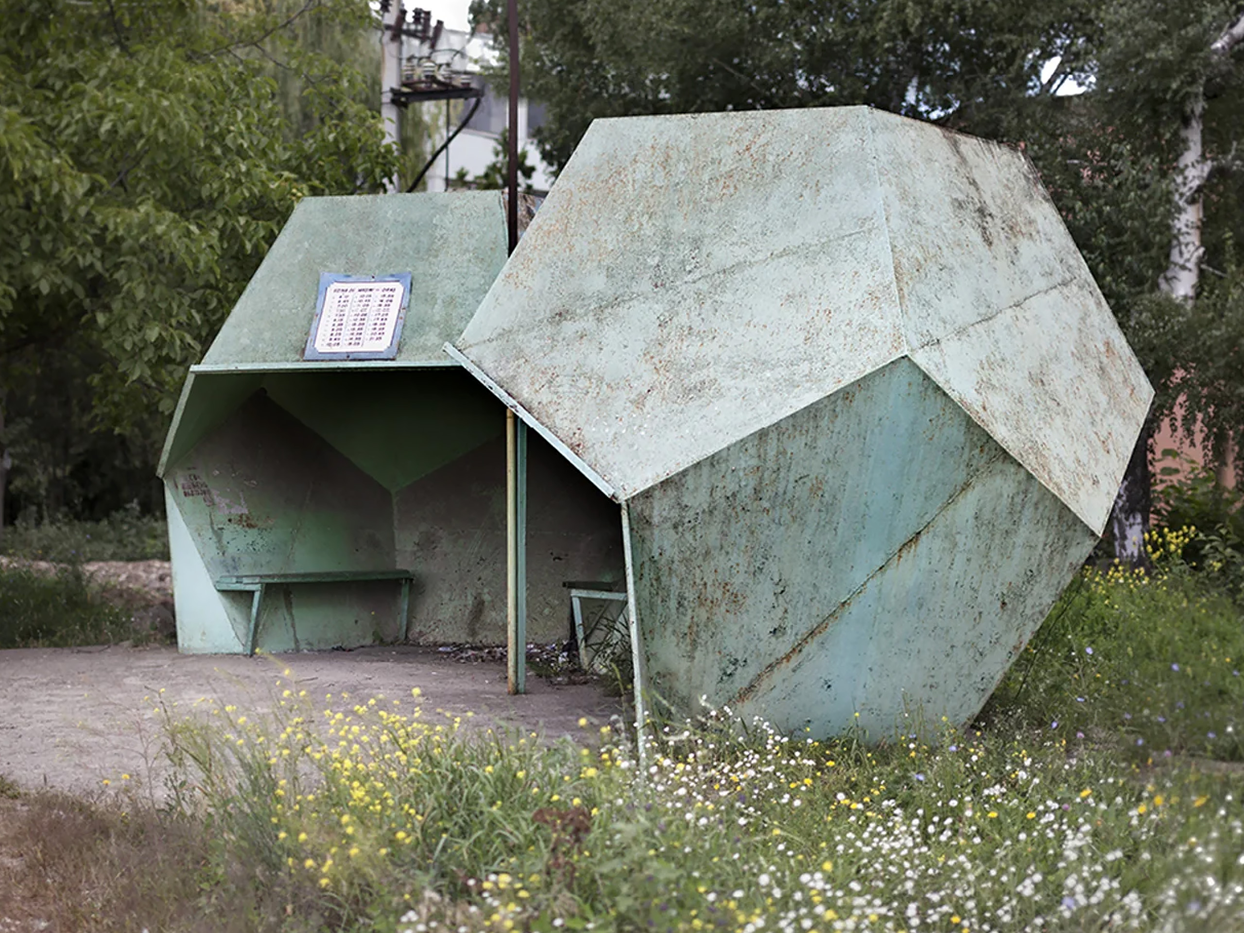Rusted green geometric sculpture in a park, surrounded by tall grasses and wildflowers.