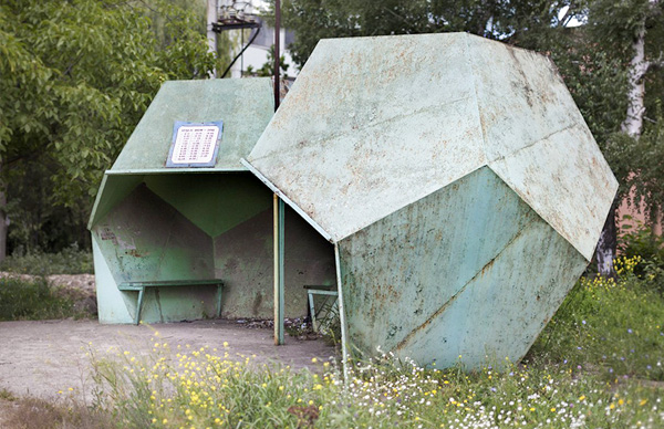 Weathered turquoise geometric metal shelter with a bench inside, set in a grassy park.