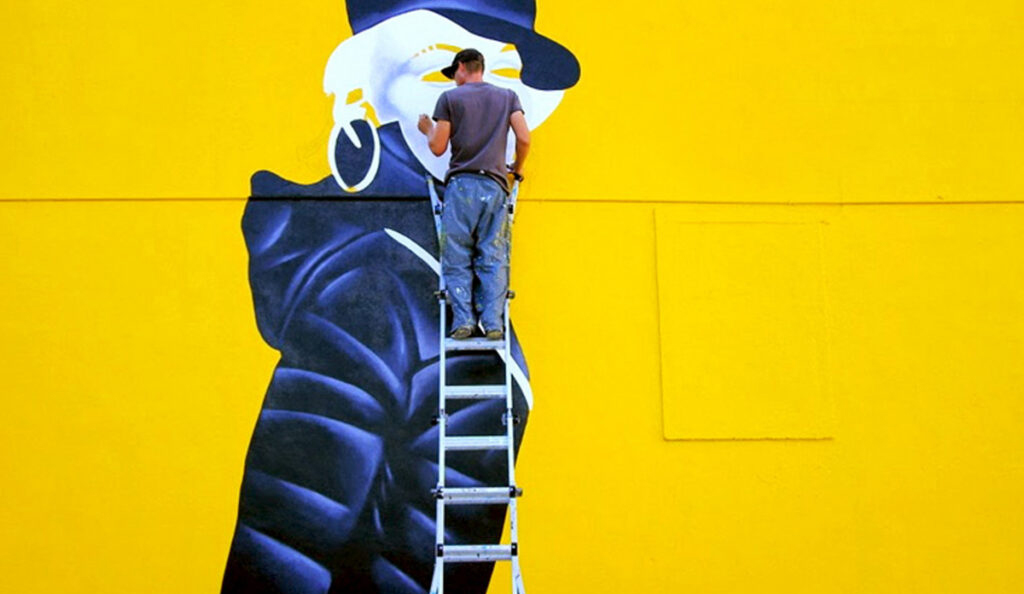 A painter on a ladder adds final touches to a large mural of a dark-clothed figure on a bright yellow wall.