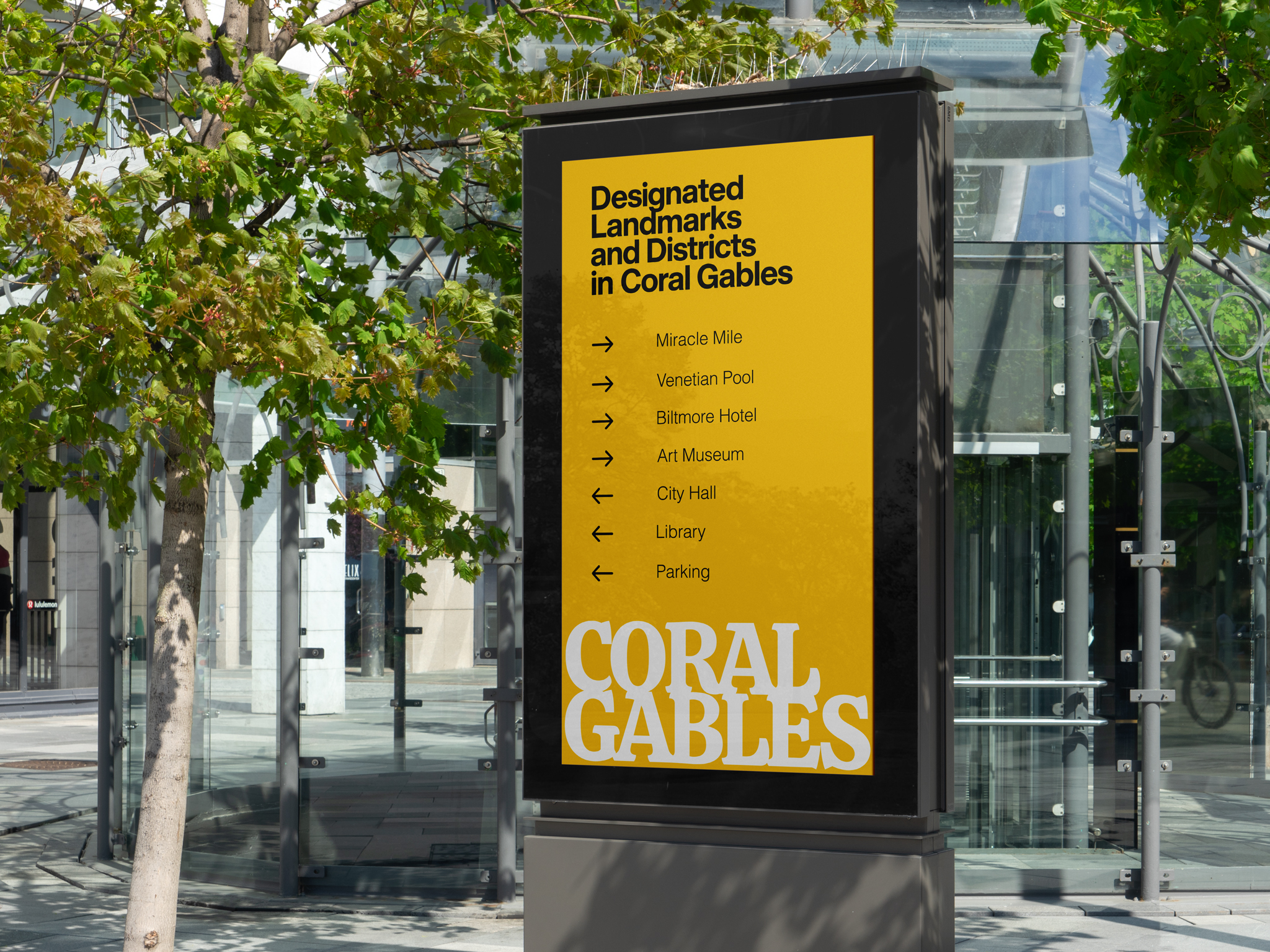 Yellow signboard listing designated landmarks and districts in Coral Gables with arrows to destinations like Miracle Mile and Venetian Pool, beside a glass building.