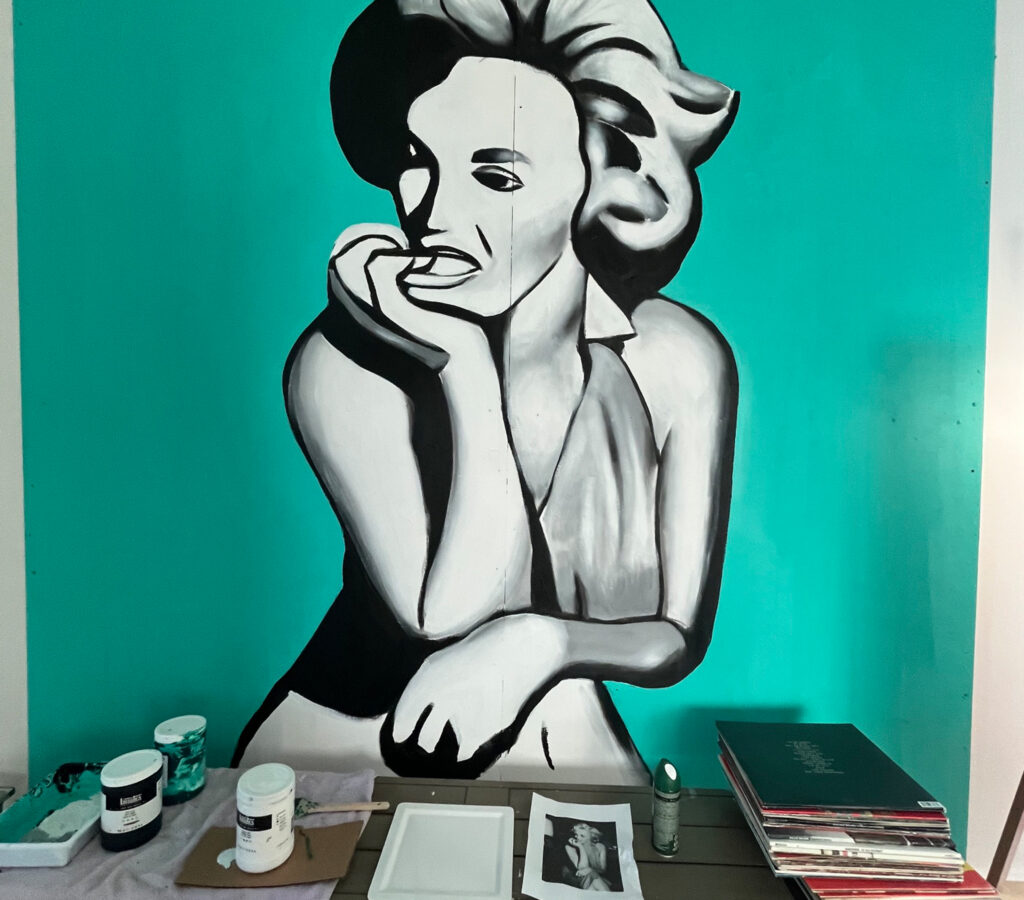 Artist's workspace with a large grayscale portrait on a teal wall; paint jars, brushes, and papers scattered on the table in the foreground.